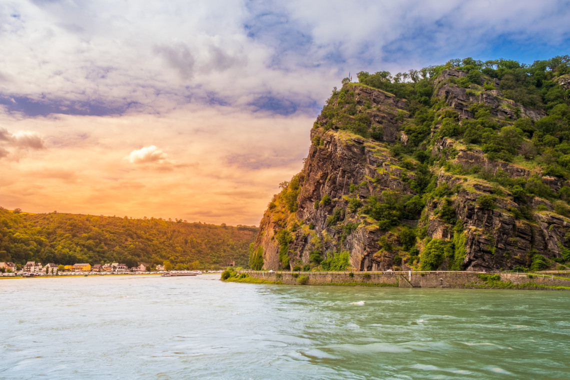 View of the steep Loreley Rock rising beside the Rhine River, with rugged cliffs, green vegetation and the river flowing through the narrow valley.