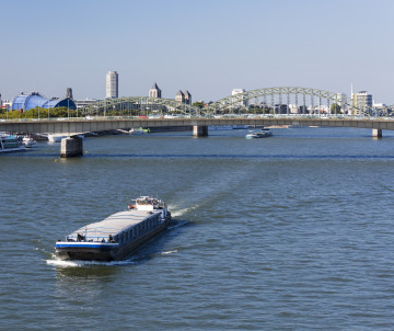 River cruise ship sailing along the Rhine in Cologne, with bridges and the city skyline extending along the riverbanks.