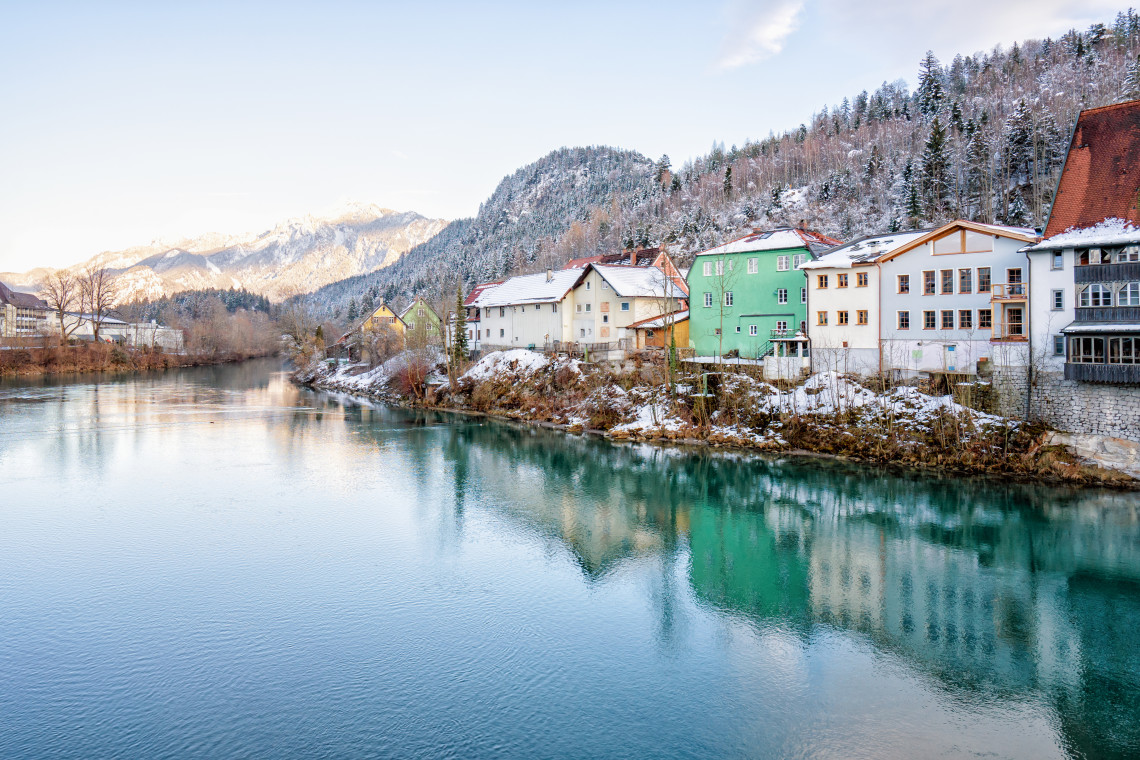 Snow-covered buildings along the turquoise Lech River in F&uuml;ssen, with forested hills and the Bavarian Alps in the background.