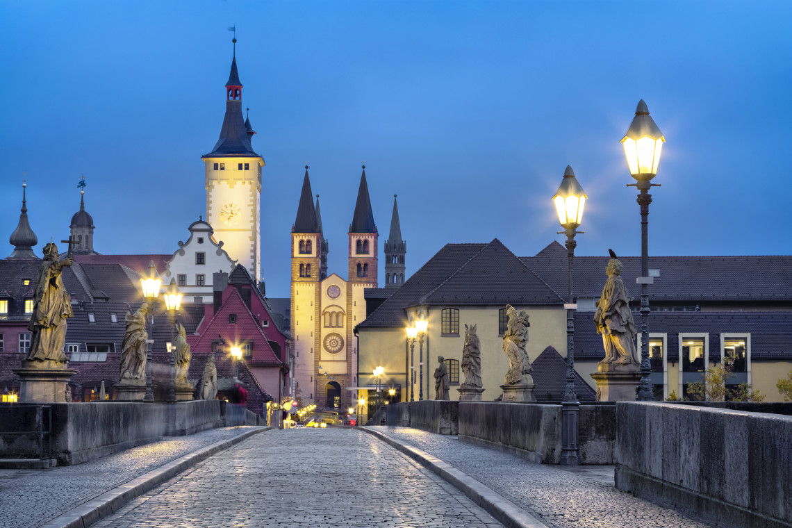 Historic Alte Mainbr&uuml;cke in W&uuml;rzburg at dusk, lined with stone statues and leading toward the illuminated towers of the old town.
