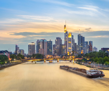 Frankfurt’s modern skyline illuminated at sunset above the Main River, with a riverboat cruising toward the city center.