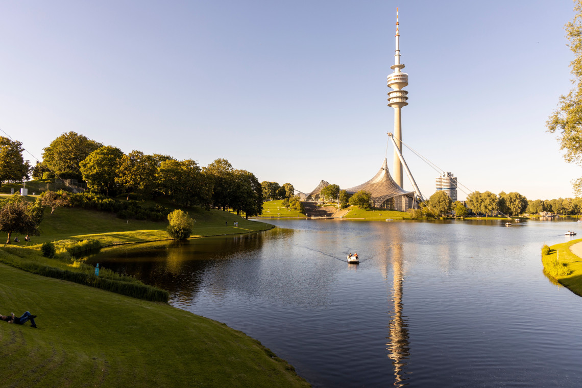 The Olympic Tower and tent-roofed sports complex rise beside the lake at Olympiapark in Munich, Germany. Green lawns and trees surround the water under a clear evening sky.