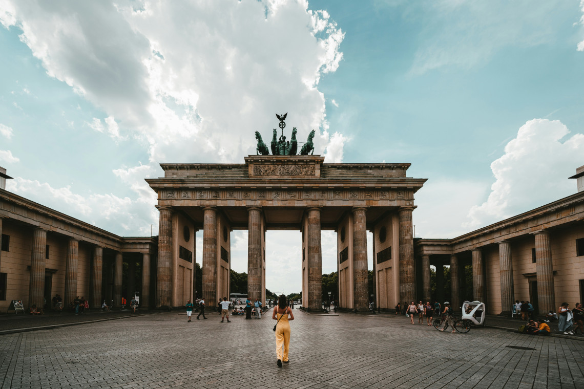 A visitor walks towards the Brandenburg Gate at Pariser Platz in Berlin, Germany. The landmark&rsquo;s sandstone columns and Quadriga sculpture are framed by a bright, cloud-filled sky.