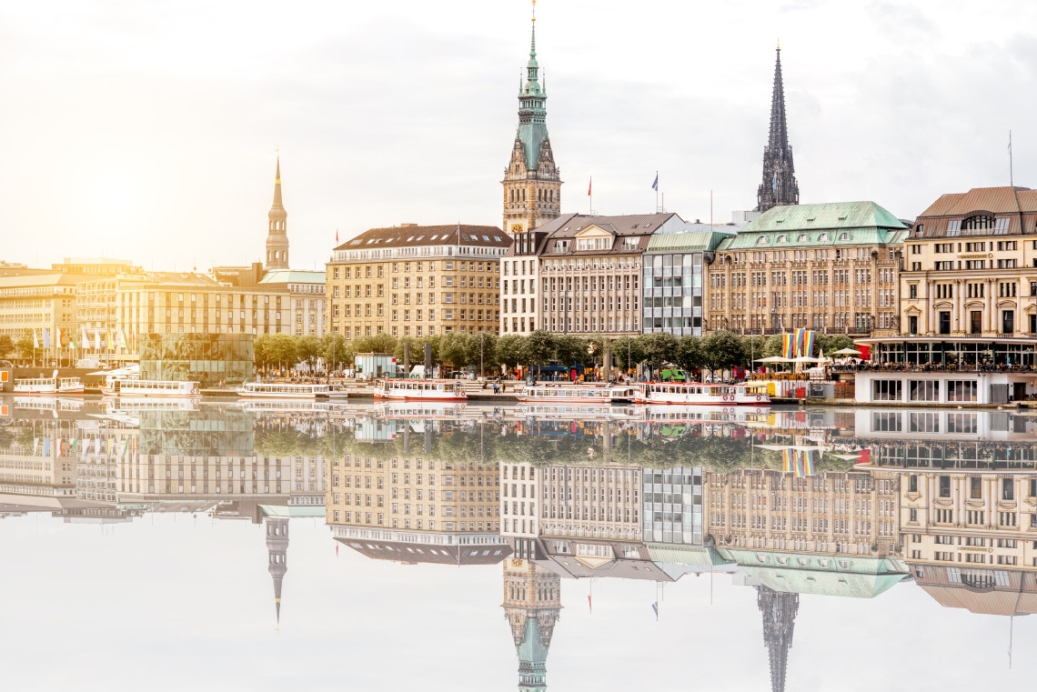 Historic buildings and church towers in central Hamburg, Germany, are reflected in the calm waters of the Binnenalster. Passenger boats line the waterfront beneath a partly cloudy sky.