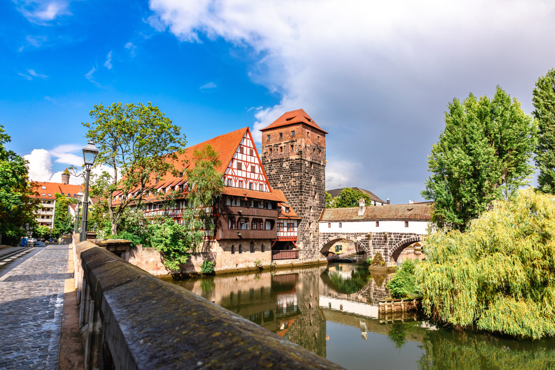 The half-timbered Weinstadel and stone Henkerturm stand beside the Pegnitz River in Nuremberg, Germany. Historic walls, a stone bridge and riverside trees are reflected in the water.