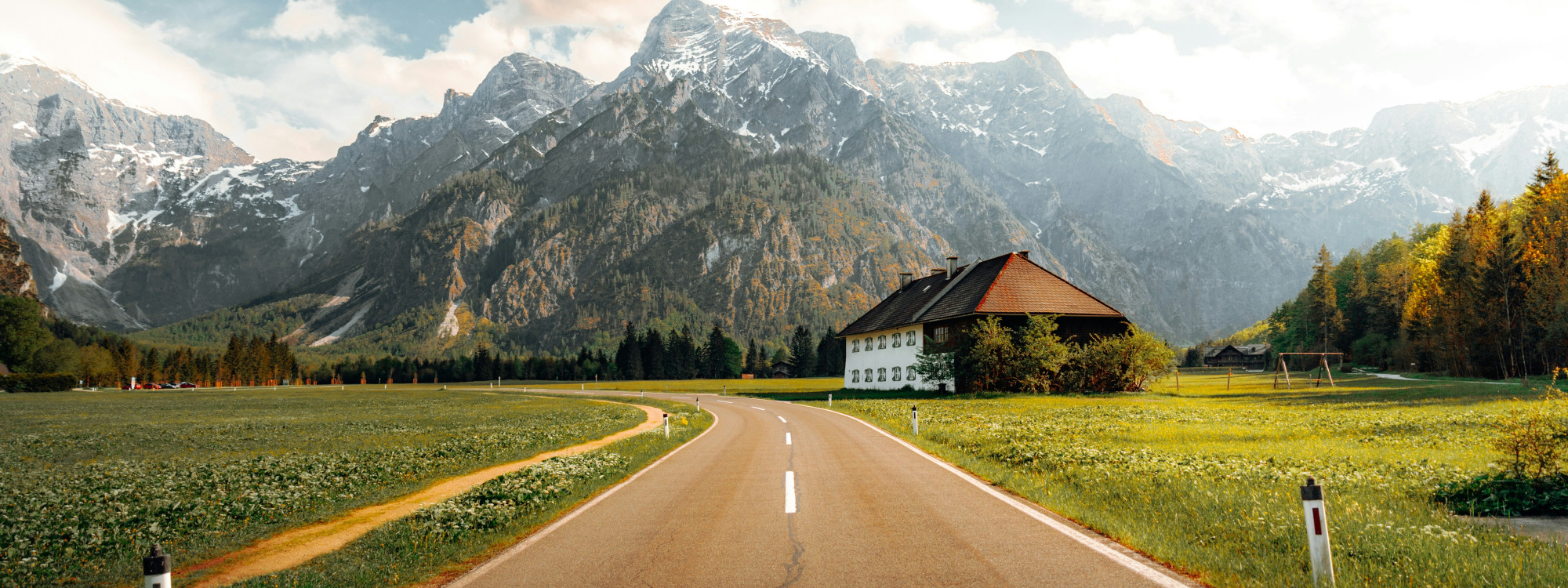 A paved scenic road crosses a rural valley in Tyrol, Austria, leading toward forested slopes and rugged Alpine mountains. A traditional farmhouse stands beside open meadows beneath a partly cloudy sky.