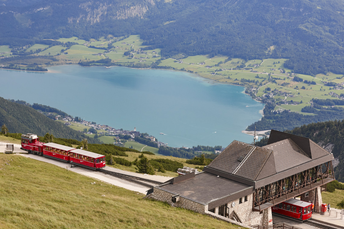 A red cogwheel train travels along the Schafberg Railway above Lake Wolfgang in Salzburg, Austria. The mountain station overlooks the blue lake, surrounding villages and forested Alpine slopes.