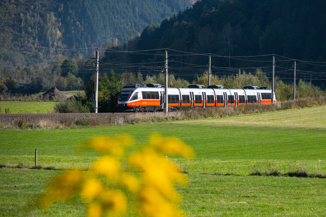 A modern regional train travels through a green Alpine valley in Austria&rsquo;s Salzkammergut region. Open fields, forested mountains and railway infrastructure surround the passenger train.