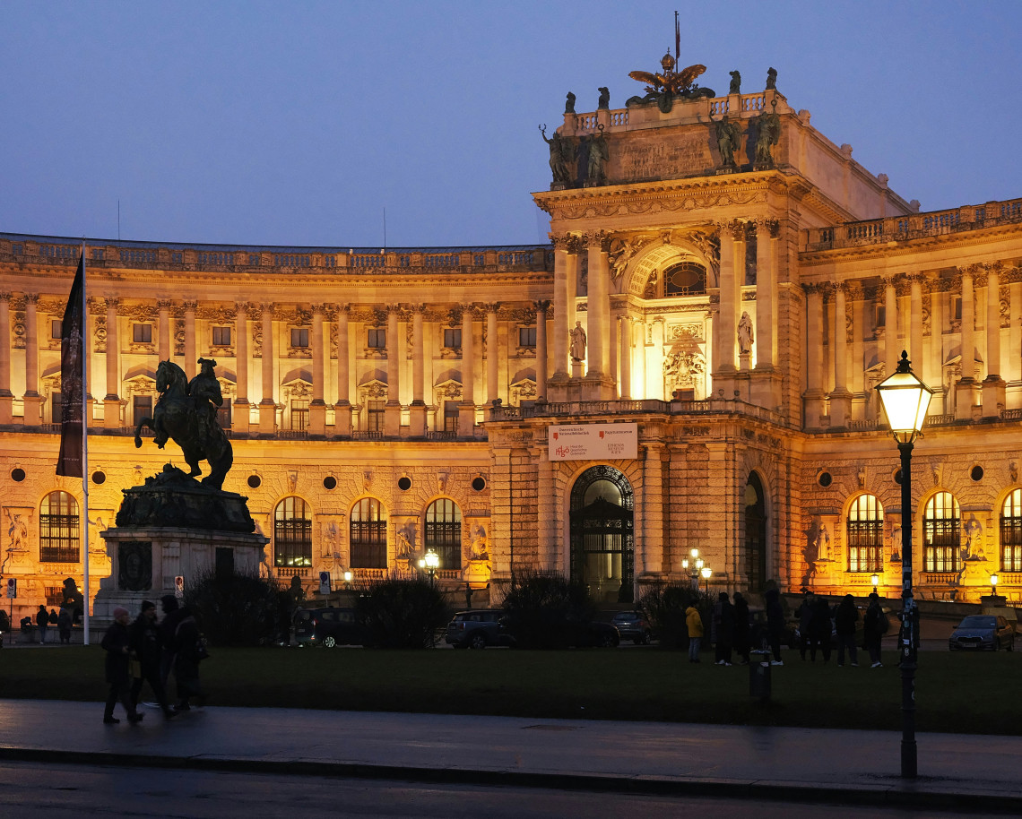 The illuminated Neue Burg wing of Hofburg Palace stands beside an equestrian monument at Heldenplatz in Vienna, Austria. Its curved fa&ccedil;ade, columns and sculpted entrance are visible against the evening sky.