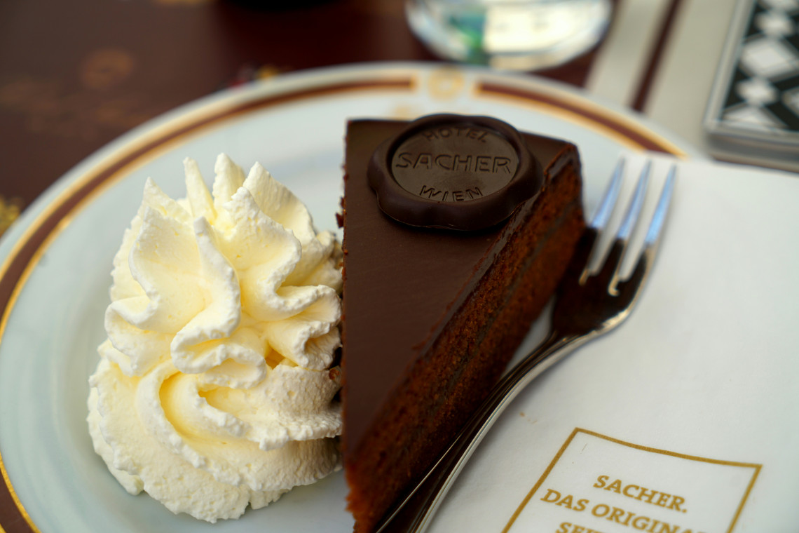 A slice of Original Sachertorte is served with whipped cream on a branded plate at Hotel Sacher in Vienna, Austria. The chocolate-glazed cake carries the Hotel Sacher seal beside a dessert fork.