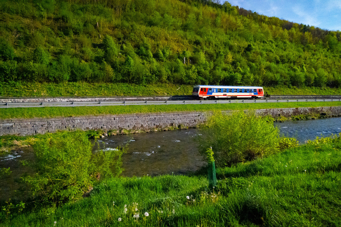 A regional passenger train travels beside a river through the green countryside of Lower Austria. Forested slopes, spring vegetation and a parallel roadway surround the railway route.