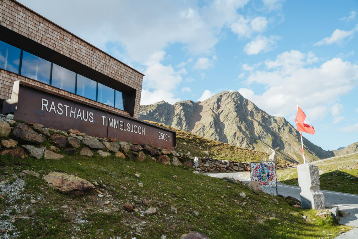 The Rasthaus Timmelsjoch stands at 2,509 metres on the Timmelsjoch High Alpine Road in Tyrol, Austria. Rocky mountain slopes, an Austrian flag and the winding pass road surround the high-altitude building.