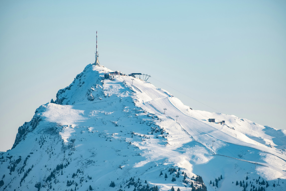 The snow-covered summit of Kitzb&uuml;heler Horn rises above Kitzb&uuml;hel, Austria, with ski lifts, mountain facilities and a communications tower visible along the ridge.