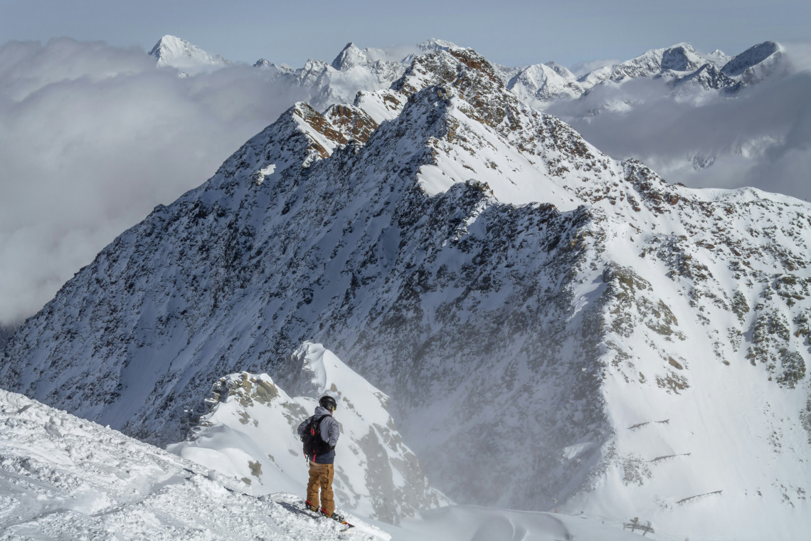 A skier stands below a steep snow-covered ridge at Rettenbach Glacier in S&ouml;lden, Austria. Rocky summits and clouds surround the high-altitude glacier landscape.