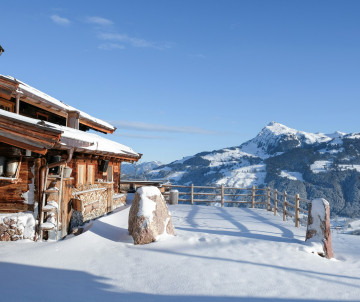 A traditional wooden chalet stands in a snow-covered mountain landscape in Tyrol, Austria. A fenced terrace overlooks forested slopes and Alpine peaks beneath a clear blue winter sky.