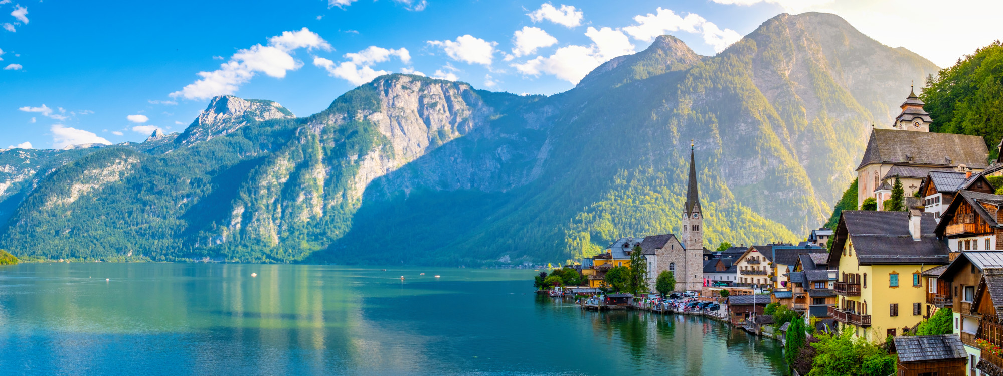 Panoramic view of Hallstatt beside Lake Hallstatt in Austria, with the village church, waterfront houses and surrounding Alpine mountains reflected in the clear water. The lake extends across the Salzkammergut landscape beneath a blue sky.