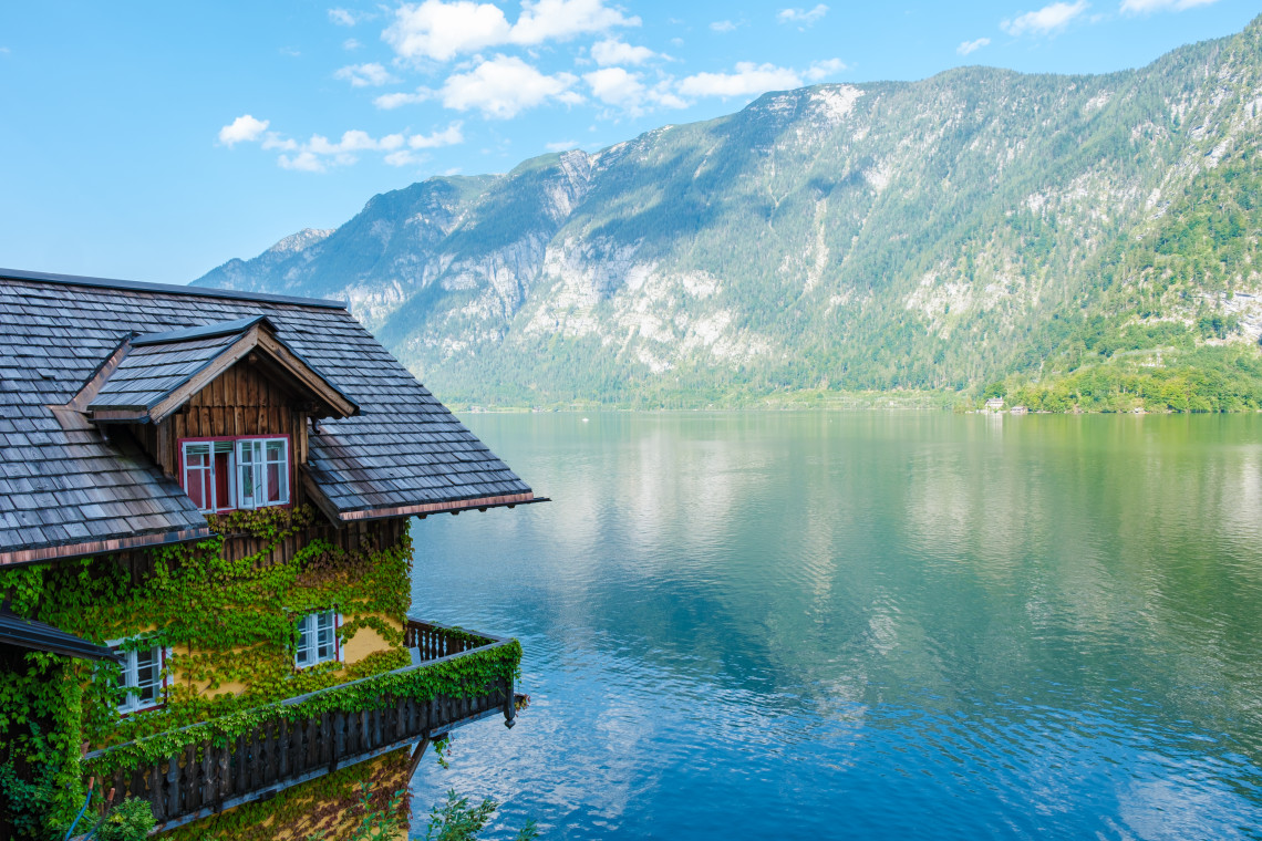 A traditional wooden house covered in greenery overlooks Lake Hallstatt in Hallstatt, Austria. Calm water and forested limestone mountains extend across the Salzkammergut landscape.