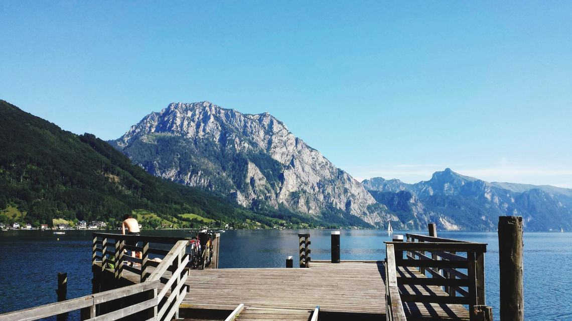 A wooden pier extends into Lake Traunsee near Gmunden, Austria, with the limestone face of Traunstein Mountain rising across the water. Clear skies and calm conditions define the Alpine lake scene.