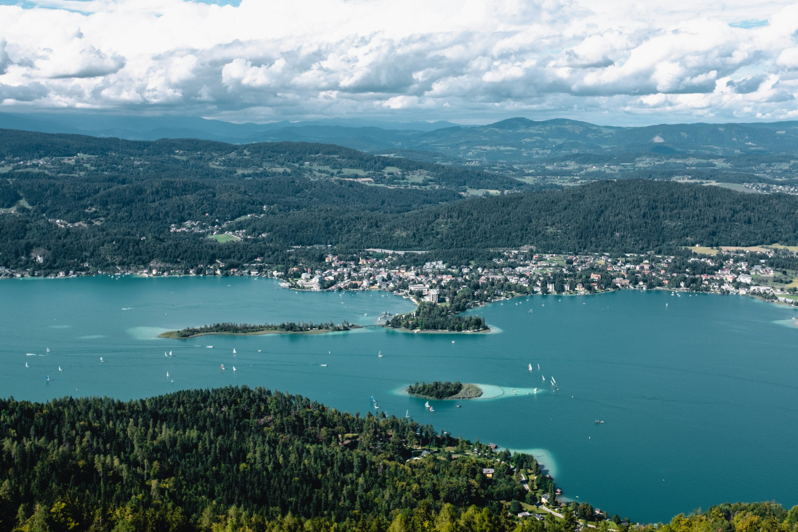 Aerial view of Lake W&ouml;rthersee in Carinthia, Austria, showing wooded islands, sailing boats and lakeside settlements. Forested hills surround the turquoise water beneath a cloudy sky.