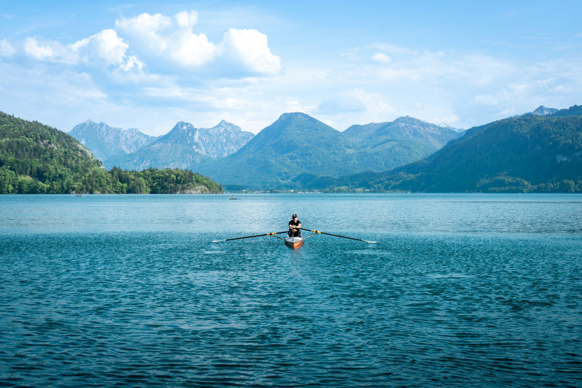 A single rower crosses Lake Wolfgang in Austria&rsquo;s Salzkammergut region. Forested mountain ranges surround the open blue water beneath a partly cloudy sky.