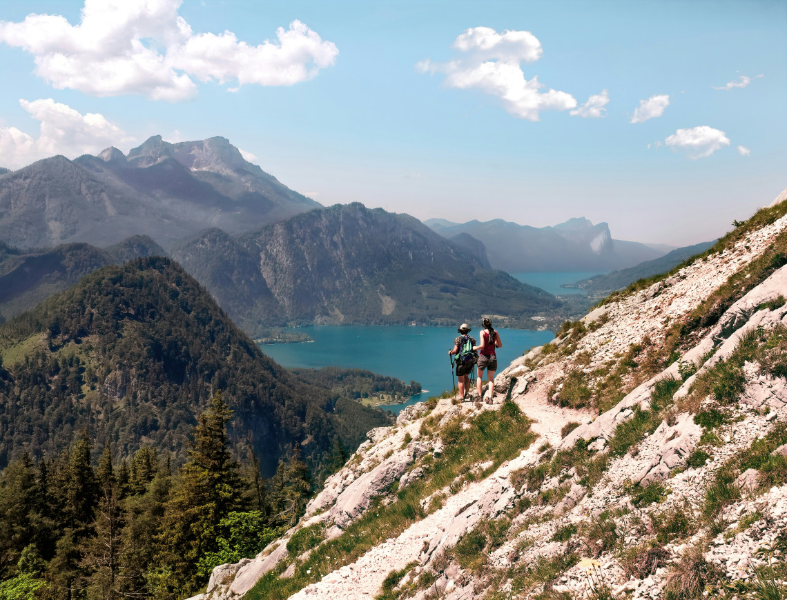 Two hikers follow a rocky trail on Schafberg in Austria&rsquo;s Salzkammergut region, with Lake Wolfgang visible far below. Steep limestone slopes, forested valleys and mountain ridges extend across the landscape.