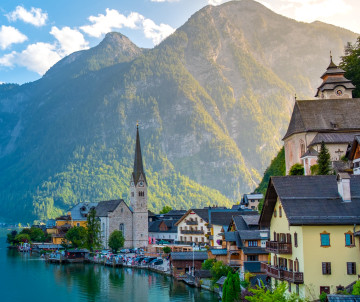 Hallstatt village in Austria sits beside Lake Hallstatt, with its historic church spire and traditional Alpine houses beneath steep forested mountains. Calm water and waterfront buildings define the lakeside setting.