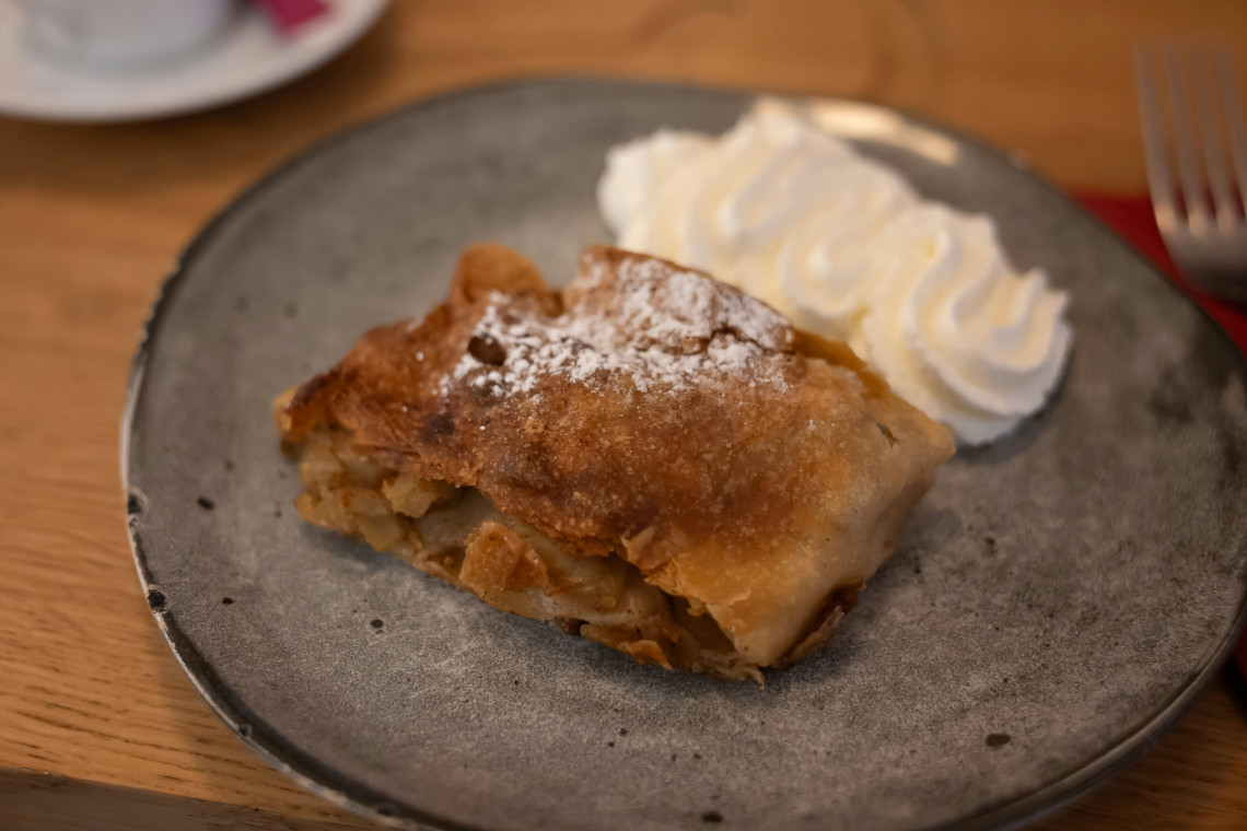 A slice of Austrian Apfelstrudel is served in Vienna, Austria, with apple filling, powdered sugar and whipped cream. The layered pastry rests on a grey ceramic plate.