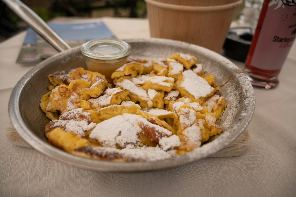 Traditional Austrian Kaiserschmarrn served in a metal pan with shredded caramelised pancake pieces and powdered sugar. A small jar of fruit compote accompanies the dish.