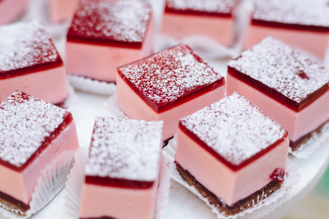Small pink Punschkrapferl cakes are displayed in Vienna, Austria, with red icing layers and a dusting of powdered sugar. Each square sponge cake is served in an individual paper case.