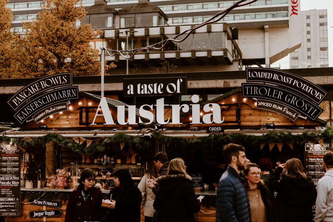 Visitors gather around an Austrian food stall at a Christmas market in Vienna, Austria. Signs advertise Kaiserschmarrn, Tiroler Gr&ouml;stl, bread bowls and other traditional Austrian dishes.
