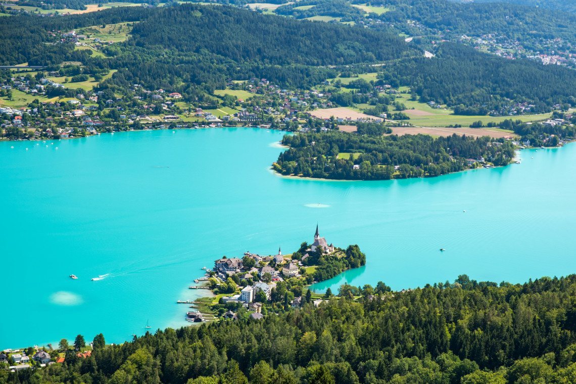 Aerial view of the Maria W&ouml;rth peninsula extending into Lake W&ouml;rthersee in Carinthia, Austria. Turquoise water surrounds the lakeside village, church and forested shoreline.