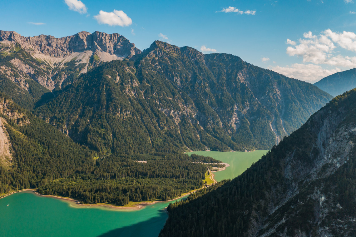 Elevated view of Plansee in Tyrol, Austria, with green-blue water winding through a steep Alpine valley. Dense forests and rocky mountain slopes surround the lake.