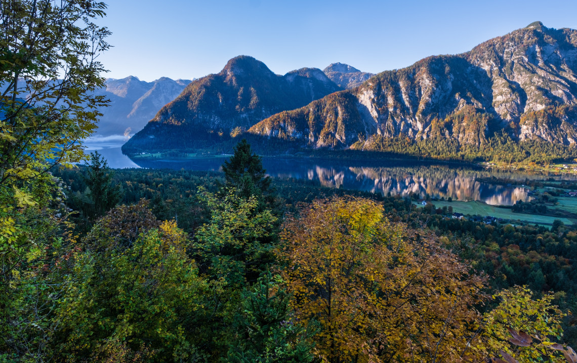 Panoramic view of Lake Hallstatt near Hallstatt, Austria, surrounded by steep forested mountains reflected in the calm water. Autumn foliage fills the foreground beneath a clear blue sky.