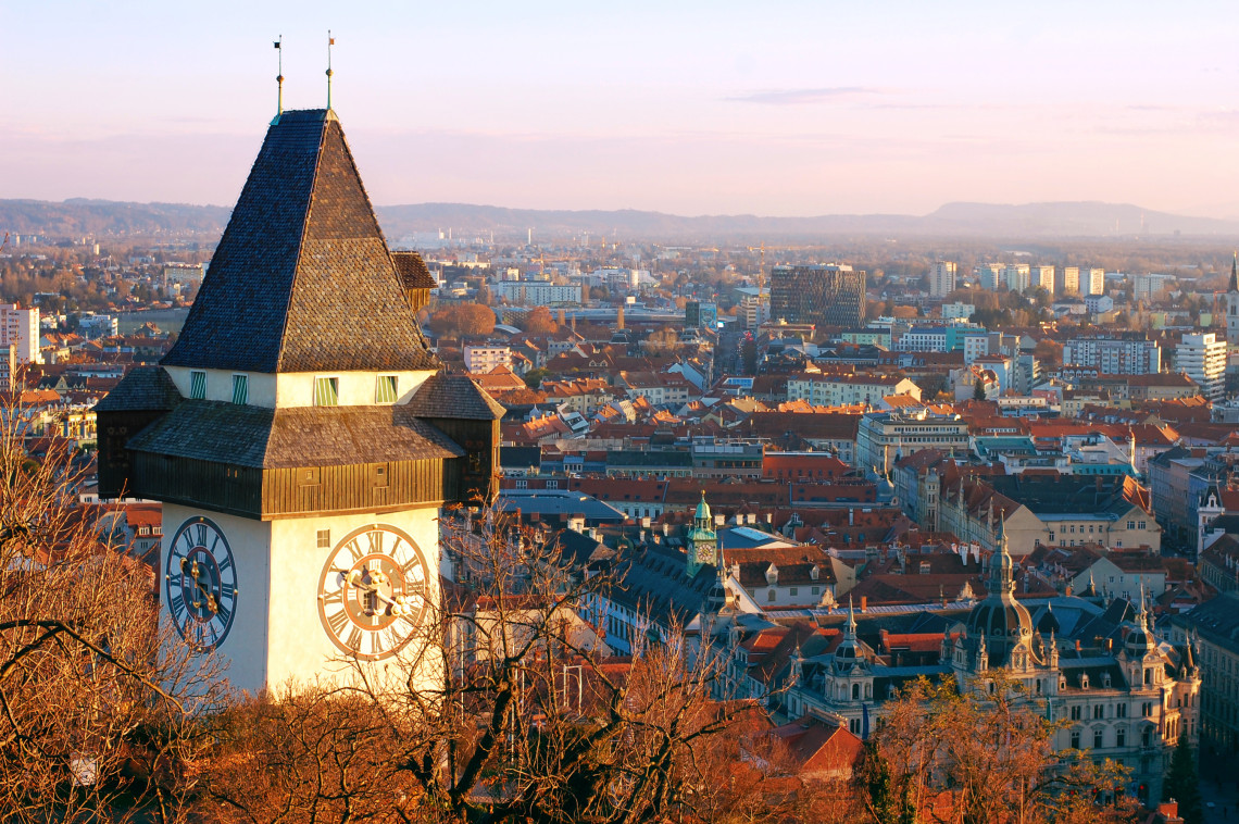 The Graz Clock Tower stands on Schlossberg above the historic centre of Graz, Austria. Red rooftops, church domes and modern city buildings extend across the landscape in warm evening light.