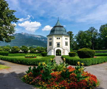 A white garden pavilion with a dark green domed roof stands among manicured hedges and colourful flowerbeds in Salzburg, Austria. Forested Alpine mountains rise behind the landscaped grounds.