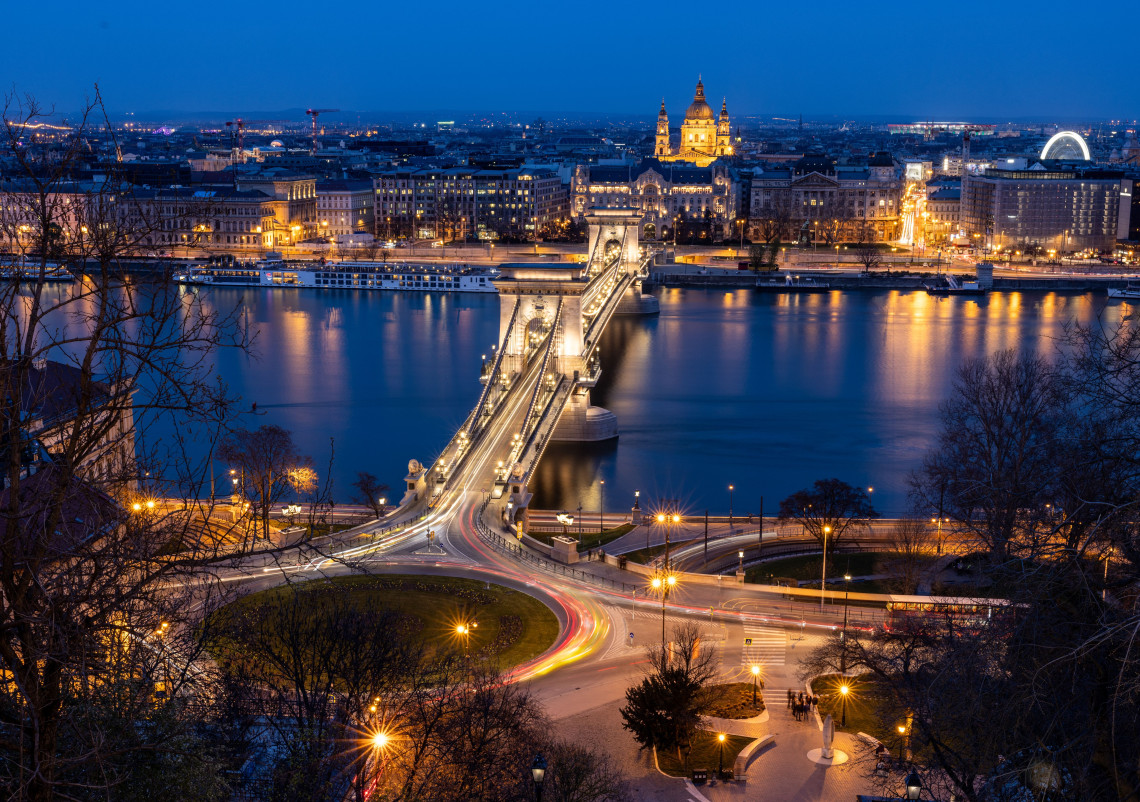 Evening view of the Sz&eacute;chenyi Chain Bridge crossing the Danube River in Budapest, Hungary. St Stephen&rsquo;s Basilica and the illuminated buildings of Pest appear beyond the bridge.