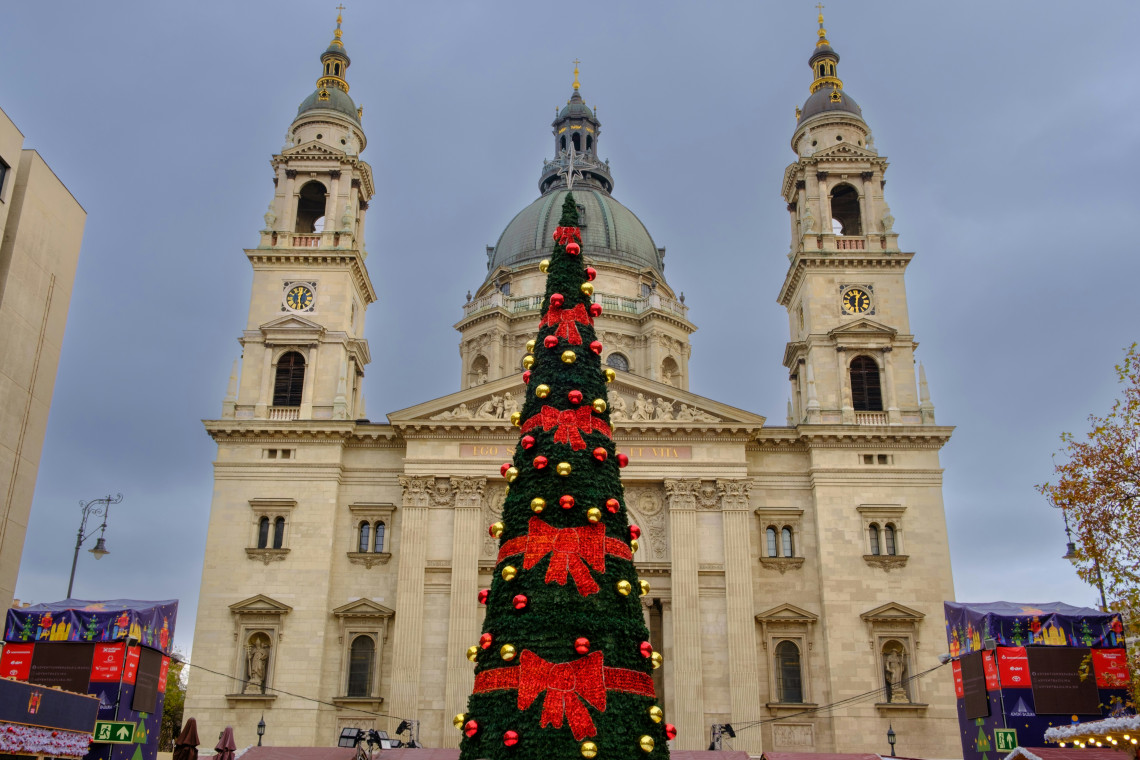 A tall Christmas tree decorated with red bows and gold ornaments stands in front of St Stephen&rsquo;s Basilica in Budapest, Hungary. The basilica&rsquo;s dome and twin bell towers frame the seasonal display.