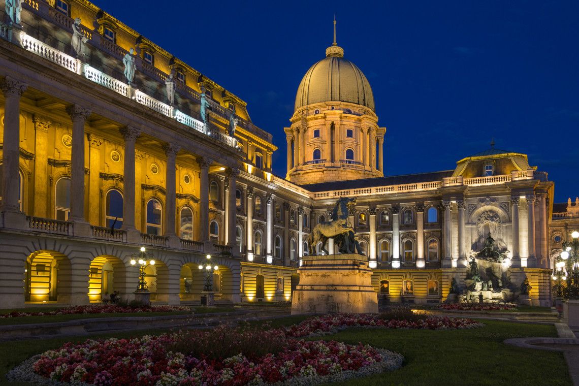 The Hungarian National Gallery at Buda Castle in Budapest, Hungary, is illuminated against the evening sky. The palace dome, equestrian statue, landscaped gardens and historic fa&ccedil;ades surround the courtyard.