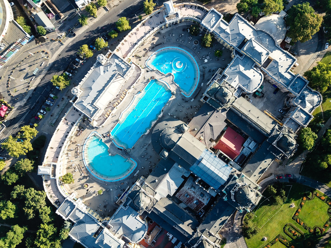 Aerial view of Sz&eacute;chenyi Thermal Bath in Budapest, Hungary, showing its three outdoor pools surrounded by Neo-Baroque buildings. Trees and landscaped grounds border the historic spa complex in City Park.