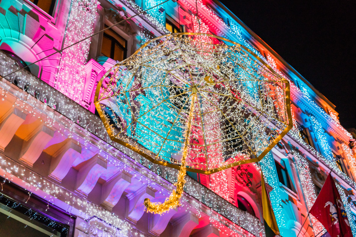 A large illuminated umbrella decoration hangs outside a historic building in Budapest, Hungary. Multicoloured fa&ccedil;ade lighting and dense strands of white lights create a seasonal display at night.