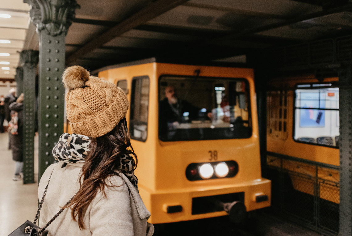 A passenger waits as a yellow train arrives on Budapest&rsquo;s Millennium Underground Railway in Hungary. The historic metro station features dark metal columns and a compact underground platform.