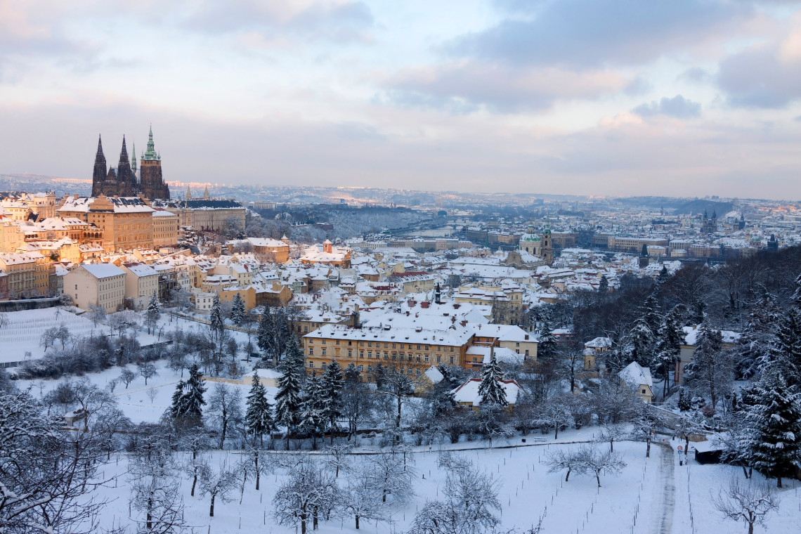 Winter panorama of Prague, Czech Republic, with snow covering rooftops, gardens and wooded hills below Prague Castle. The towers of St Vitus Cathedral rise above the historic city beneath a cloudy sky.
