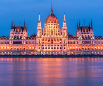 The Hungarian Parliament Building is illuminated beside the Danube River in Budapest, Hungary. Its central dome, Gothic spires and warm lights are reflected across the water at dusk.