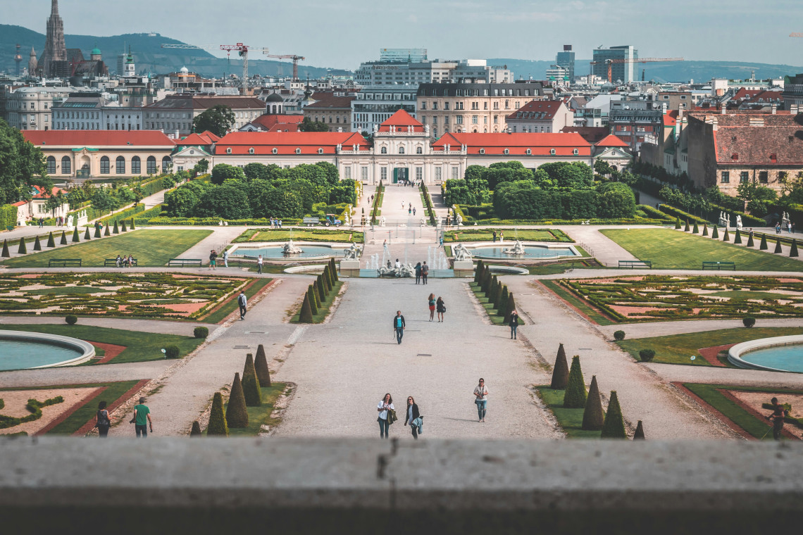 View across the formal Belvedere Gardens in Vienna, Austria, with symmetrical flowerbeds, fountains and sculpted hedges leading toward Lower Belvedere Palace. Vienna&rsquo;s city skyline rises beyond the landscaped grounds.