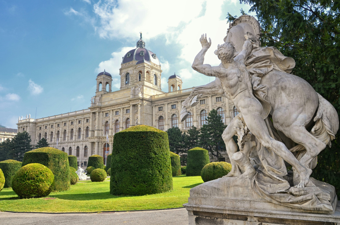 The Kunsthistorisches Museum in Vienna, Austria, stands behind manicured hedges and a large stone sculpture in Maria-Theresien-Platz. Its domed roof and detailed Neo-Renaissance fa&ccedil;ade rise beneath a blue sky.