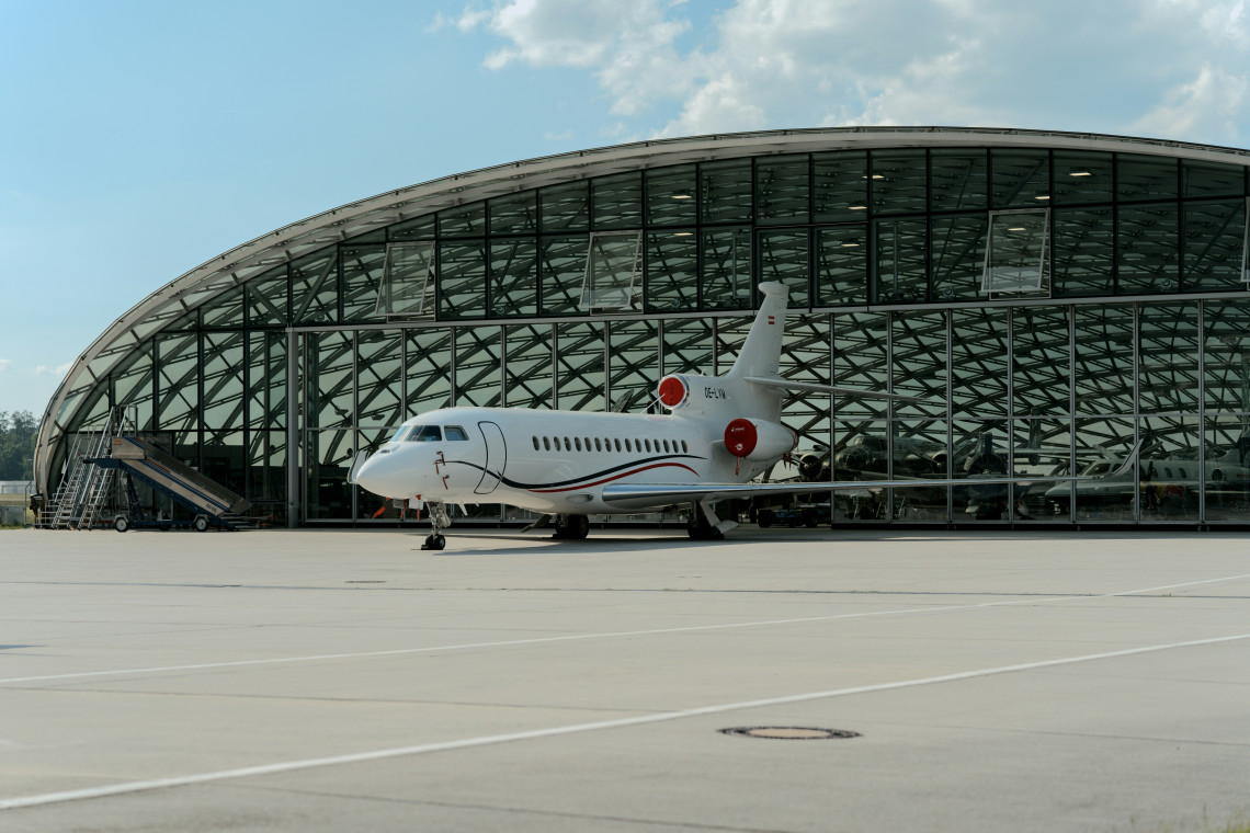 A private jet is parked outside Hangar-7 in Salzburg, Austria. The aircraft stands before the landmark&rsquo;s curved glass-and-steel structure beneath a clear blue sky.