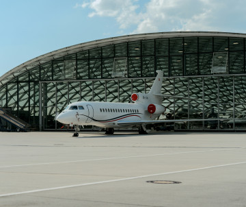 A private jet is parked on the apron outside Salzburg Airport in Salzburg, Austria. The aircraft stands in front of the airport’s curved glass terminal under a partly cloudy sky.