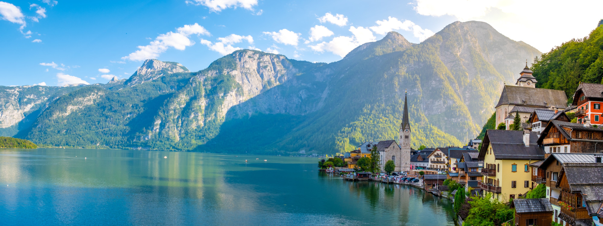 Panoramic view of Hallstatt beside Lake Hallstatt in Austria, with the village church, waterfront houses and steep Alpine mountains reflected in the clear water.