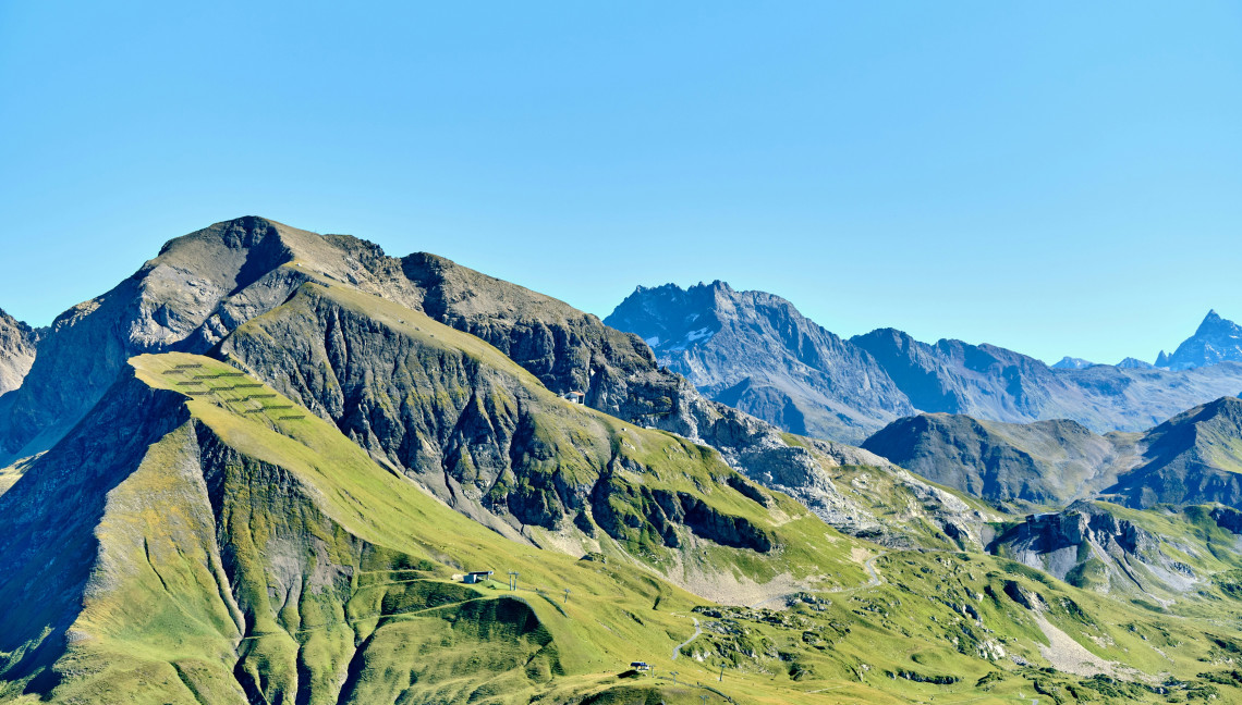 Mountain ridges and green Alpine slopes surround Lech am Arlberg in Vorarlberg, Austria. Hiking paths and small mountain structures cross the high-altitude landscape beneath a clear blue sky.