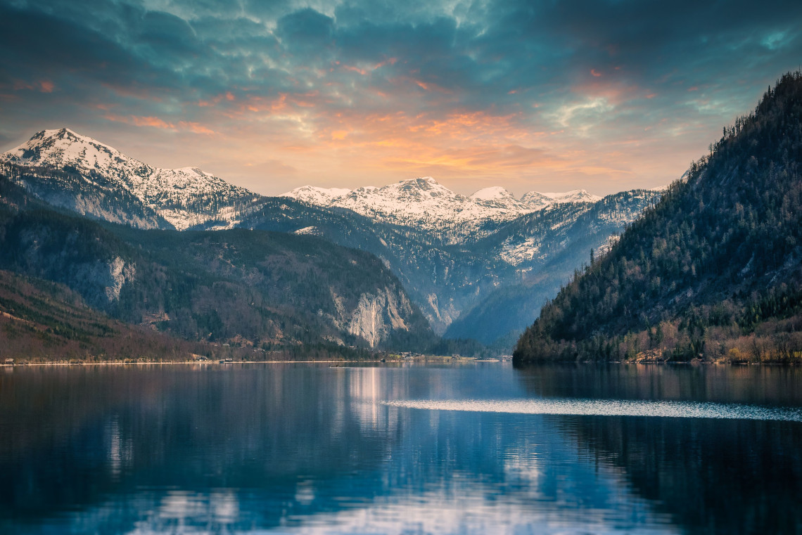 Grundlsee in Styria, Austria, reflects forested slopes and snow-covered mountains beneath a warm evening sky. The long Alpine lake extends through the Salzkammergut landscape.